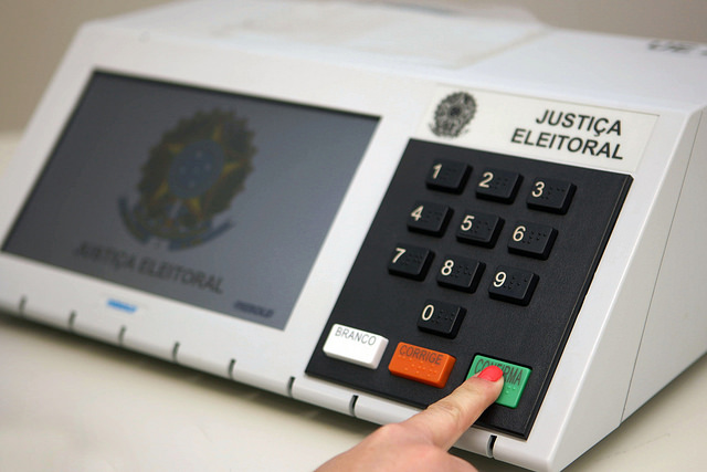 Foto de urna eletrônica eleitoral, com um dedo de uma mulher pressionando a tecla verde Confirma.