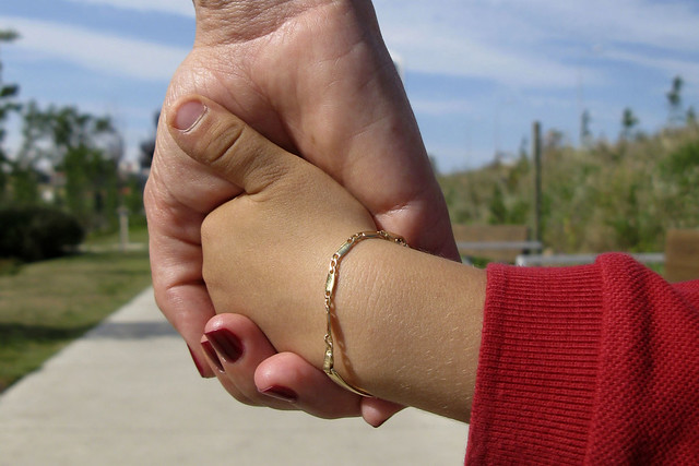 Foto mostra o detalhe da mão de uma mulher segurando a mão de uma criança em um parque.