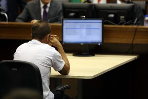 Foto mostra um homem sentado em uma sala de audiência em um tribunal durante realização de audiência de custódia.