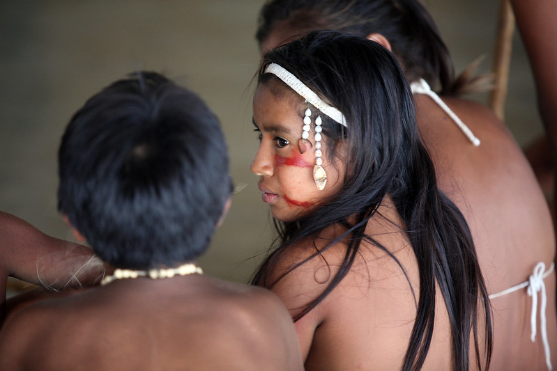 Foto mostra duas meninas e um menino indígena sentados, de costas na foto.