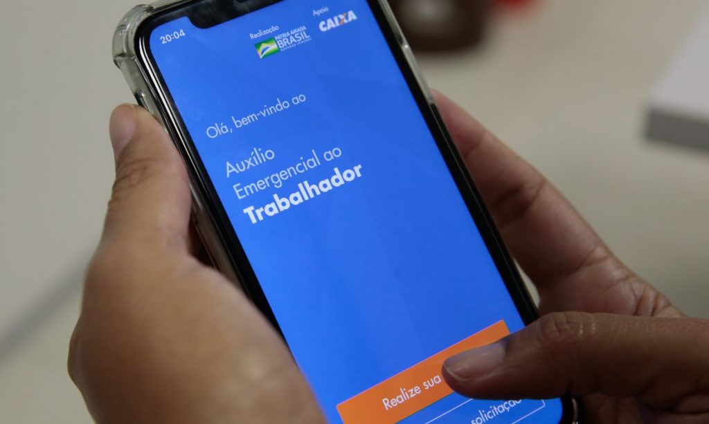 Foto de mão segurando um cellular que está acessando o aplicativo Auxílio Emergencial