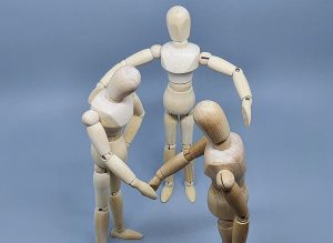 Foto ilustrativa sobre conciliação, com três bonecos de madeira articulados, sendo que dois estão fazendo movimento de aperto de mãos e o terceiro está com os braços abertos em volta de ambos, como se estivessem firmando um acordo