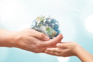 Dois pares de mãos de pessoas brancas, uma de pessoa adulta, a outra de uma criança, enquanto seguram juntas uma miniatura do planeta Terra.