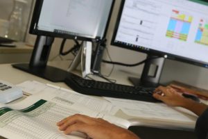 Foto mostra a mão de uma pessoa em cima de uma folha de papel com uma tabela enquanto a outra digita no teclado de um computador, como se estivesse verificando uma informação.