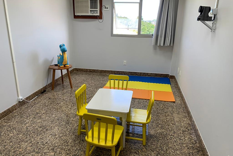 Foto de sala de depoimento especial mostra na parede, ao fundo, aparelho de ar-condicionado e uma janela. A sala tem como mobiliário uma mesa pequena, para crianças, com 4 cadeiras amarelas também pequenas. Atrás, tem um tapete colorido para a criança ficar no chão.