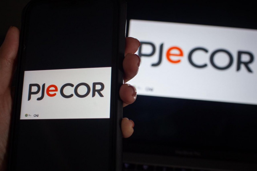 Foto mostra uma mão segurando um celular e na tela tem a logomarca do PJeCOR. Ao fundo, tem um monitor de computador com a mesma logomarca.