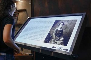 Foto de mulher acessando a exposição virtual em tela interativa.