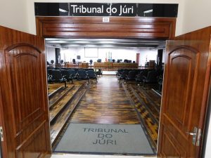 Imagem das portas de entrada para o Tribunal do Júri, em primeiro plano, acima, descrição da sala “ TRIBUNAL DO JÚRI 03”, em segundo plano a plateia e ao fundo o júri, compostas somente por cadeiras.