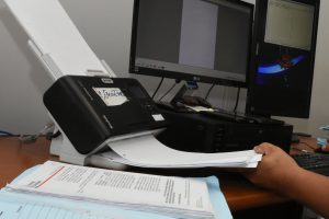Foto de uma estação de trabalho, com monitor ao fundo. É possível ver as mãos de uma pessoa digitalizando uma folha de processo.