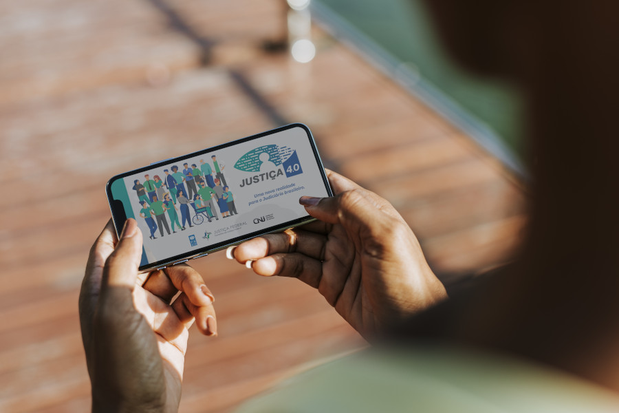 Foto mostra as mãos de uma mulher negra segurando um celular. Na tela, se vê a logomarca do programa Justiça 4.0.