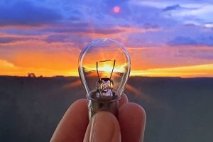 Foto mostra os dedos de um homens segurando uma lâmpada pequena, em um cenário amplo, onde se pode ver ao fundo o nascer do sol.
