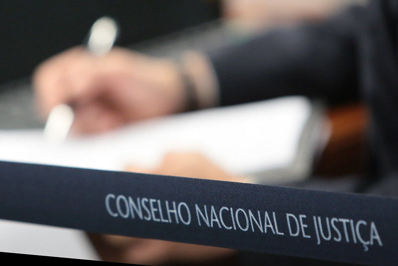 Foto mostra um homem escrevendo ao fundo, desfocado. Em destaque uma faixa onde se lê "Conselho Nacional de Justiça".