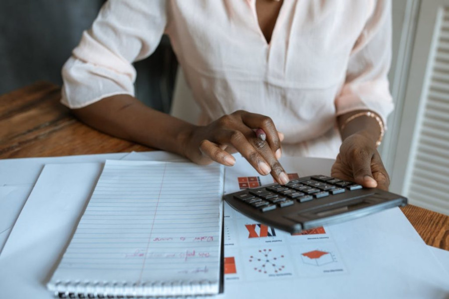 Foto mostra uma mulher negra usando uma calculadora em cima de uma mesa cheia de papeis.