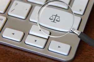 Foto mostra um teclado de computador com uma lupa em uma tecla onde se vê a balança símbolo da Justiça.
