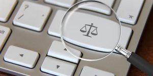 Foto mostra um teclado de computador com uma lupa em uma tecla onde se vê a balança símbolo da Justiça.