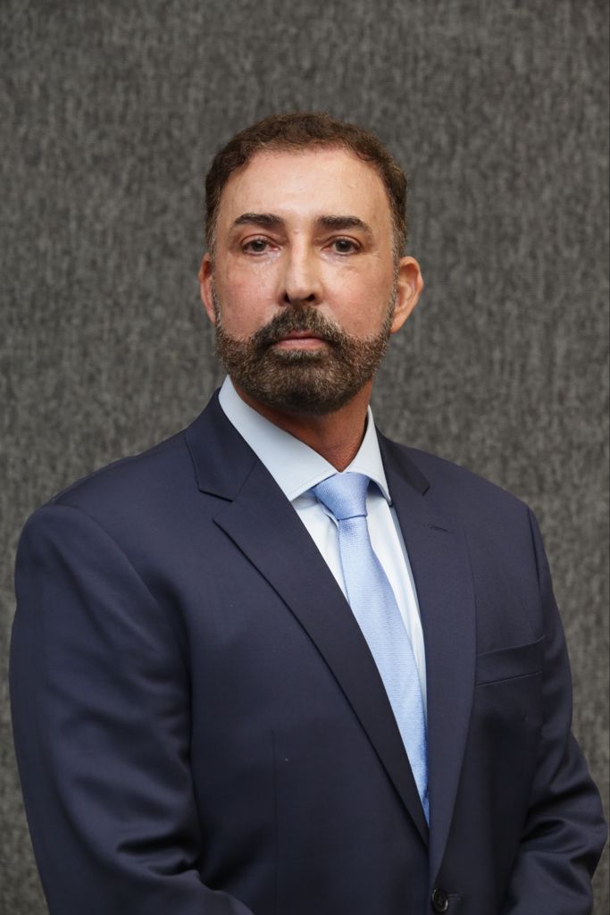 Foto do conselheiro do CNJ Sidney Pessoa Madruga. Ele está usando paletó azul escuro e camisa e gravata azul claras, em frente a um fundo texturizado cinza.