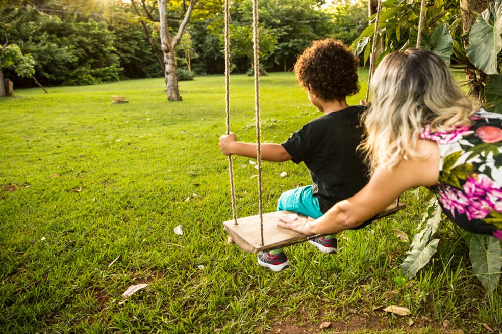 Foto mostra, em um cenário gramado e com árvores ao fundo, uma criança sentada em um balanço e uma mulher, atrás dele, controlando o balanço.