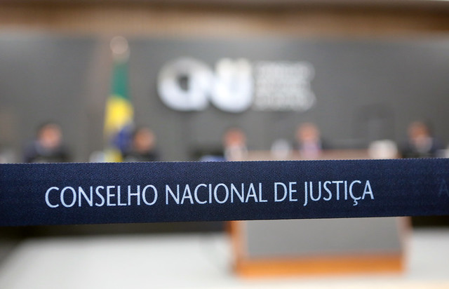 Foto do plenário do CNJ. Ao fundo, fora de foco, é possível ver pessoas sentadas no plenário durante a sessão, com a logomarca do CNJ na parede. Em destaque, faixa de divisão onde se lê "Conselho Nacional de Justiça"