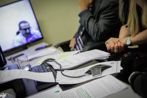 Foto mostra detalhes de um homem e uma mulher sentados em uma mesa participando de videoconferência. à frente deles, na mesa, podem se ver vários papeis e o mouse do computador.