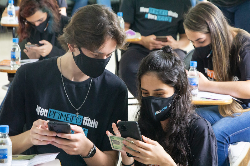 Foto de um jovem e uma jovem, segurando seus celulares e se ajudando a fazer o cadastro eleitoral. Eles estão em uma sala onde é possível ver outros jovens tamb´ em acessando o celular.