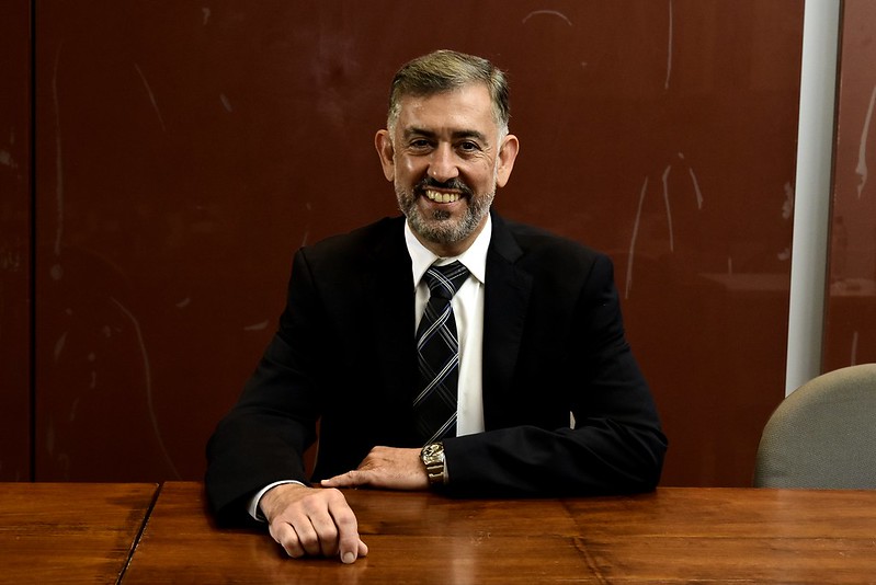 Foto mostra o desembargador sentado em uma mesa, vestindo terno e sorrindo.