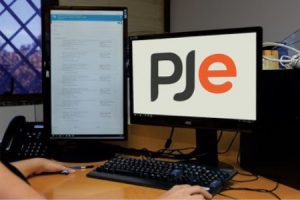 Foto mostra as mãos de uma pessoa usando um computador com dois monitores. Em um deles, está a logomarca do PJe.