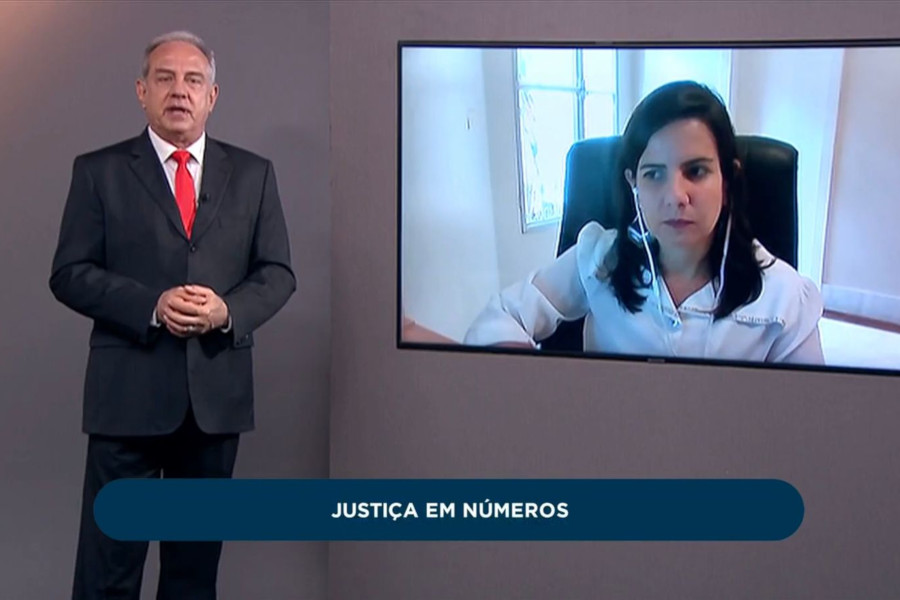 Foto mostra, no estúdio de programa, o apresentador Guilherme Menezes em pé. A seu lado, tem um telão onde se vê a diretora do Departamento de Pesquisas Judiciárias do DPJ, Gabriela Soares.