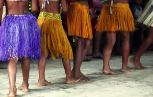 Foto de mulheres (apenas vistas da cintura para baixo) indígenas vestindo trajes típicos e descalças.