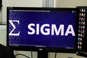 Foto mostra um monitor de computador com a logomarca do SIGMA.