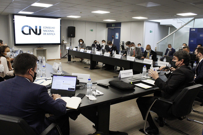 Foto geral da reunião, com as pessoas participanntes sentadas em mesa em U e, ao fundo, um telão com a logomarca do CNJ.