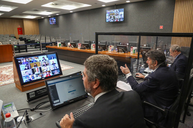 Foto no Plenário do CNJ mostra sentados, de costas para a foto, o secretário-geral do CNJ (à esquerda), o ministro Luiz Fux (ao centro) e o secretário especial de Programas do CNJ. O ministro Fux está falando e os três estão olhando para um telão no centro do Plenário, onde estão visíveis as demais pessoas que participaram por videoconferência da reunião.