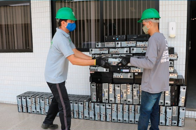 Foto mostra dois homens, usando equipamentos de proteção individual e retirando os equipamentos de informática descartados.