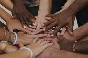 Foto mostra cerca de 10 mãos de homens e mulheres, pessoas brancas e negras, uma em cima da outra, ilustrando diversidade e equidade.