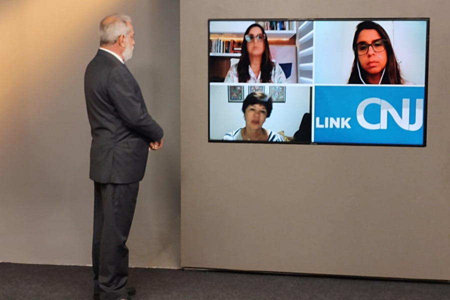 Foto mostra momento da gravação do programa no estúdio, com o apresentador Guilherme Menezes olhando para o telão na parede onde estão, por videoconferência, as pessoas participantes da edição como entrevistadas.