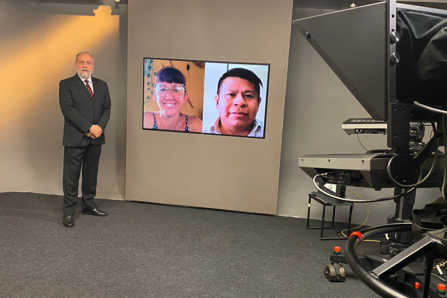 Foto mostra o apresentador Guilherme Menezes no estúdio, ao lado do telão onde se vê as duas pessoas convidadas.