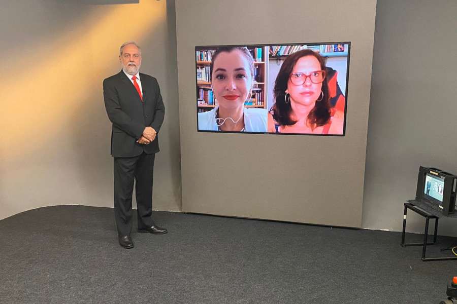 Foto mostra o apresentador Guilherme Menezes posando no estúdio ao lado do telão, em que se vê as imagens das duas entrevistadas por videoconferência.