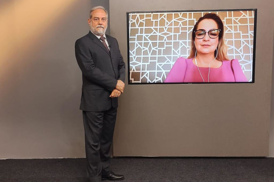 Foto mostra o apresentador no estúdio, posando ao lado do telão. No telão, está a imagem da entrevistada por videoconferência.