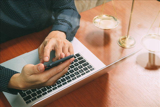 Foto mostra um homem sentado em uma mesa mexendo em um celular em frente a um notebook.