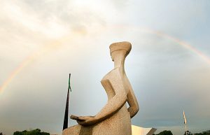 Foto da estátua em homenagem à deusa grega da Justiça Thêmis, que fica em frente ao STF, tendo ao fundo um arco-íris.