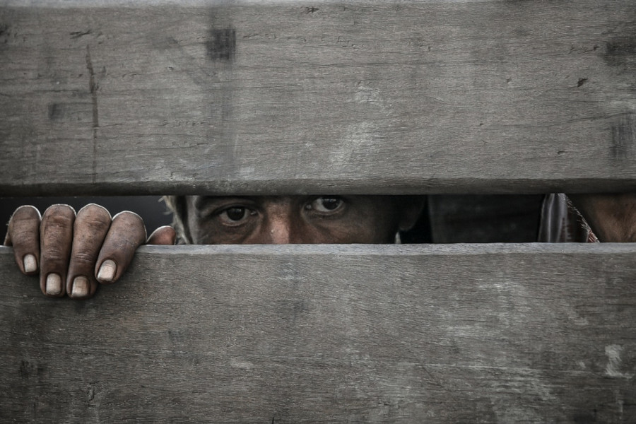 Foto mostra um homem atrás de uma cerca de madeira. É possível ver os olhos dele e uma das mãos por uma fresta.