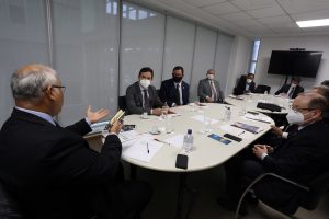 Foto mostra momento da reunião, com participantes sentados em uma sala, ao redor de uma mesa comprida.