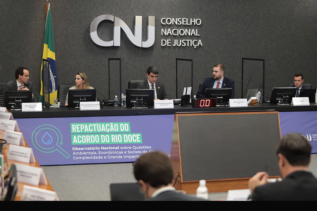 Foto mostra bancada principal do Plenário do CNJ com o conselheiro Bandeira e outras pessoas sentadas e, na frente, um banner de apoio com o texto Repactuação do Acordo do Rio Doce.
