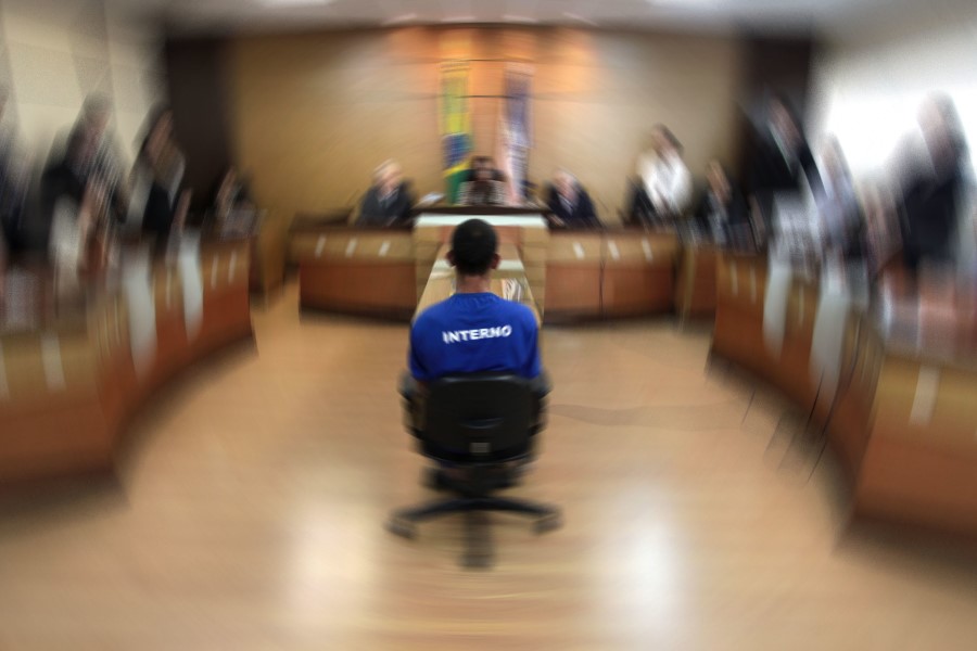 Foto de audiência de custódia realizada em sala de audiências do TJES, em 2015. A foto está com um filtro onde estão "borradas" as imagens das pessoas presentes, sendo possível ver claramente, ao centro, o homem que está sendo julgado, sentado de costas onde se lê a palavra "INTERNO" na camisa que ele usa.
