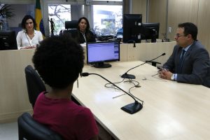 Foto mostra ambiente de audiência do Fórum, com uma mulher negra de costas sentada em uma mesa com um microfone à sua frente. Do outro lado da mesa é possível ver um homem, também com um microfone à sua frente. E à frente dos dois, em uma bancada, tem a a juíza e uma outra mulher, sentadas e olhando para os dois.