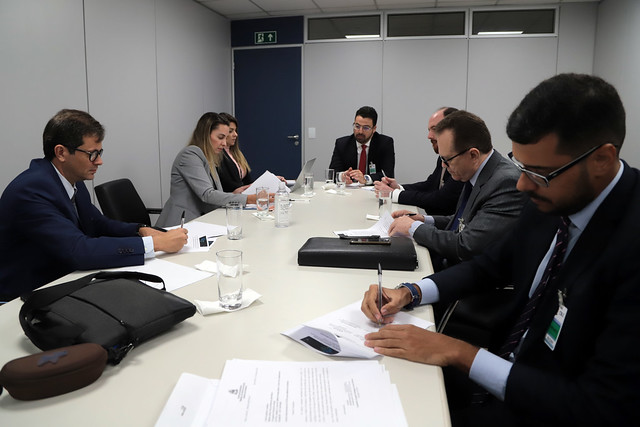 Foto mostra momento da audiência de conciliação, com sete pessoas em, uma sala sentadas em volta de uma mesa e conversando.