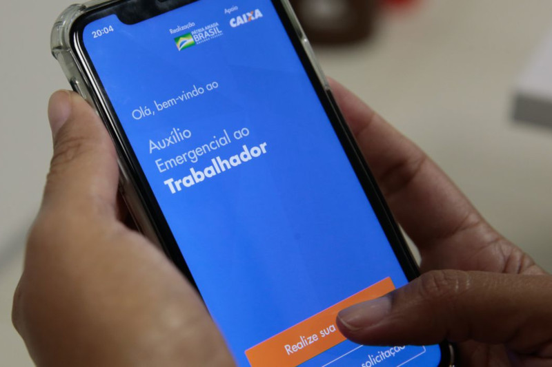 Foto de mão segurando um cellular que está acessando o aplicativo Auxílio Emergencial.