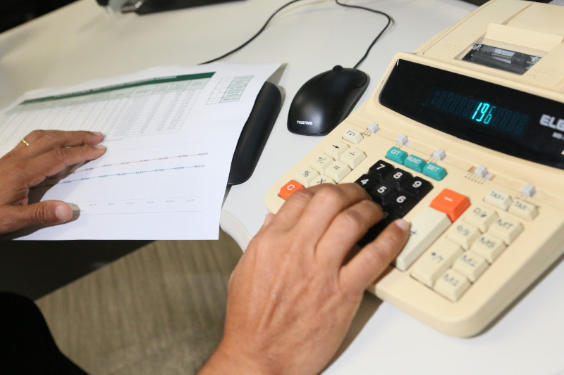 Foto mostra a mão de uma pessoas teclando uma calculadora a partir de números de uma planilha.