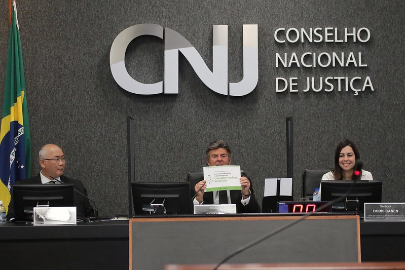 Foto mostra o ministro Luiz Fux, sentado na bancada principal do Plenário do CNJ, segurando a cartilha. À esquerda dele, na foto, está o conselheiro do CNJ Richard Pae Kim. À direita, está a juíza auxiliar da Presidência do CNJ Doris Canen.