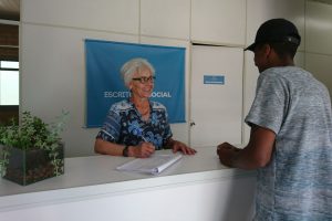 Foto mostra um homem sendo atendido por uma mulher sorridente no balcão de Escritório Social.