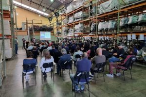 Foto mostra momento do evento, com diversas pessoas sentadas em um grande depósito e, ao fundo, um instrutor falando e apontando para um telão.